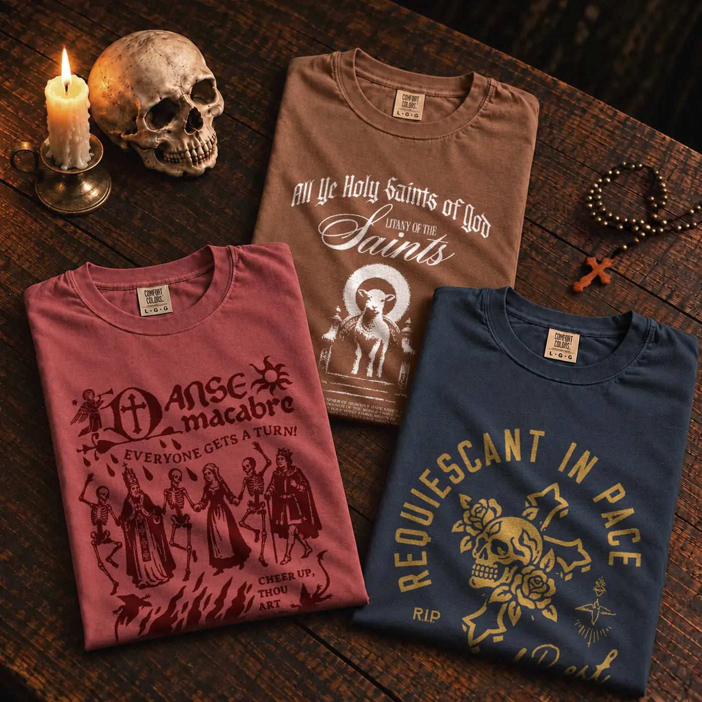 The three Days of the Dead shirts folded on a dark table with a candle, a skull, a rosary and a statue of Our Lady